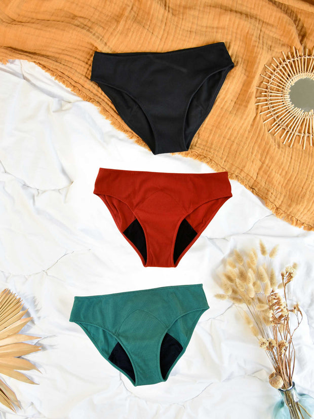 lot de 3 culottes de règles colorés , rouge, verte, noire à plat sur un lit. 